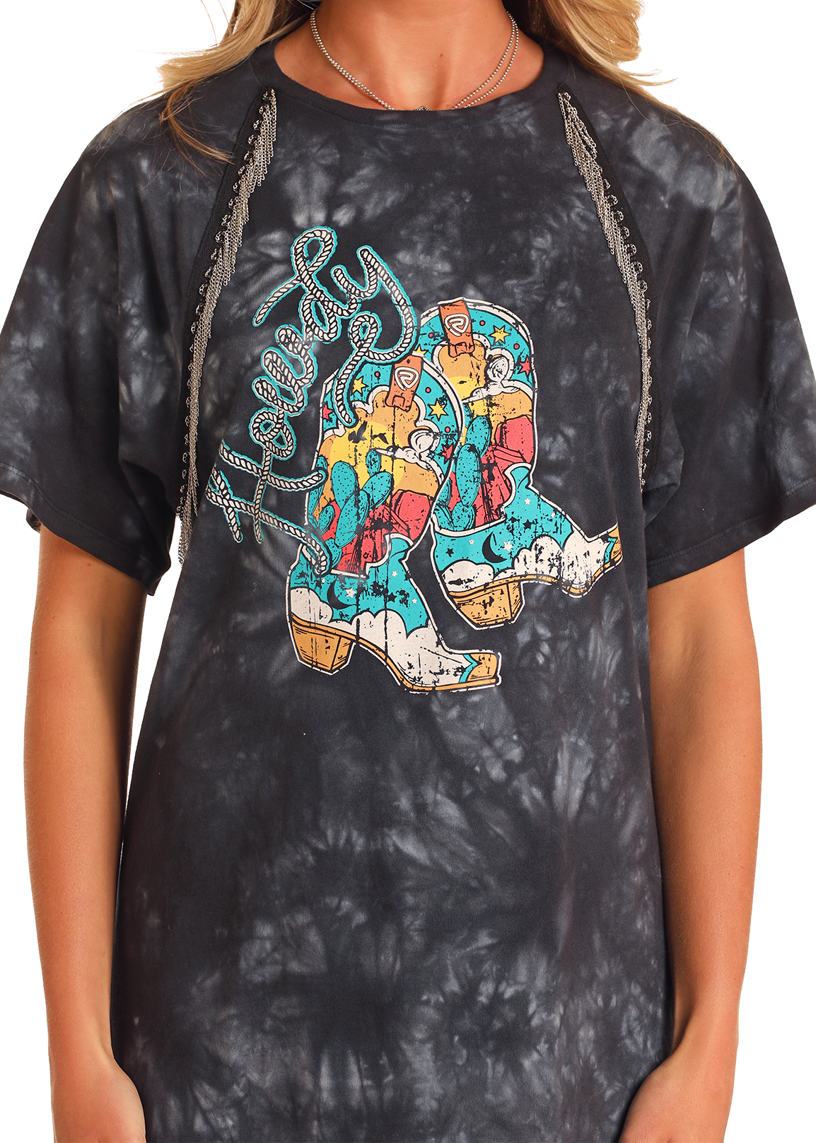 A person wears the Rock & Roll Denim Chain Fringe Graphic T-Shirt Dress, featuring a black tie-dye design, distressed colorful boot graphic with "Howdy" in turquoise script, and decorative shoulder fringe.