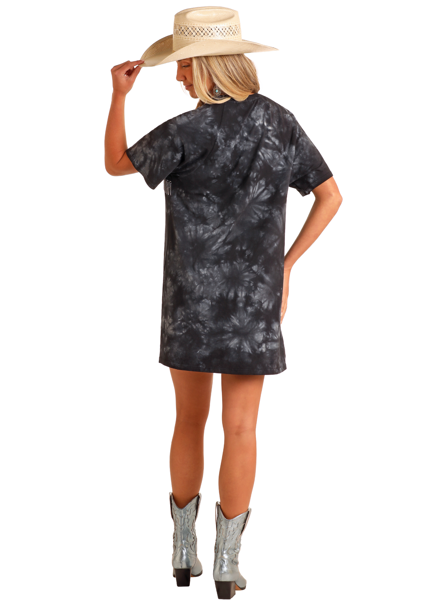 A woman faces away in a black and gray tie-dye Chain Fringe Graphic T-Shirt Dress by Rock & Roll Denim, paired with a straw hat and metallic cowboy boots. She holds her hat’s brim against a plain white background.