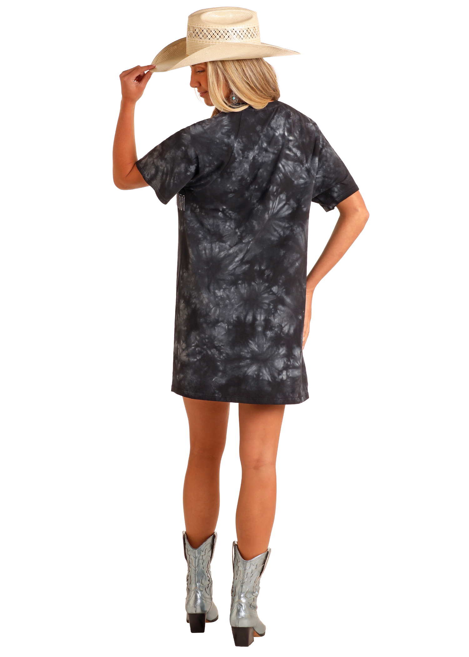 A woman faces away in a black and gray tie-dye Chain Fringe Graphic T-Shirt Dress by Rock & Roll Denim, paired with a straw hat and metallic cowboy boots. She holds her hat’s brim against a plain white background.