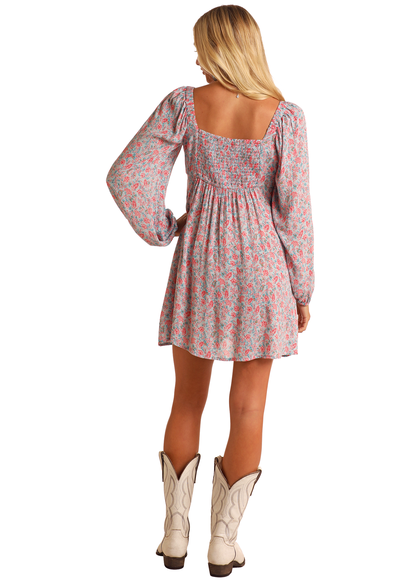 A woman with long blonde hair faces away, wearing the Rock & Roll Denim Floral Mini Dress—pink and blue with a smocked bodice and square neckline—perfect for everyday style, paired chicly with white cowboy boots.