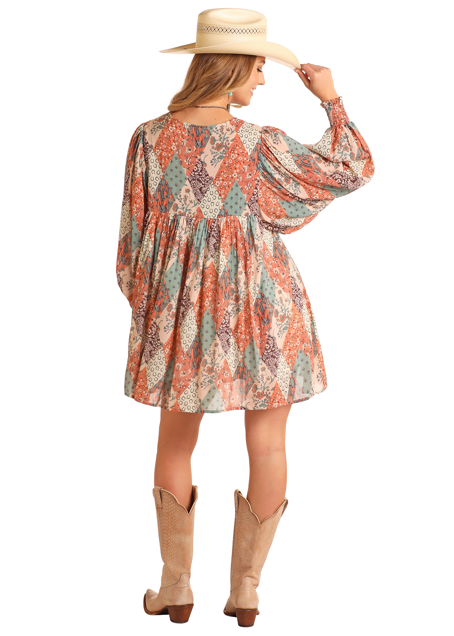 A woman in the Rock & Roll Denim Patchwork Print Mini Dress, wide-brimmed hat, and tan cowboy boots stands facing away, holding her hat’s brim—an effortlessly stylish everyday look.