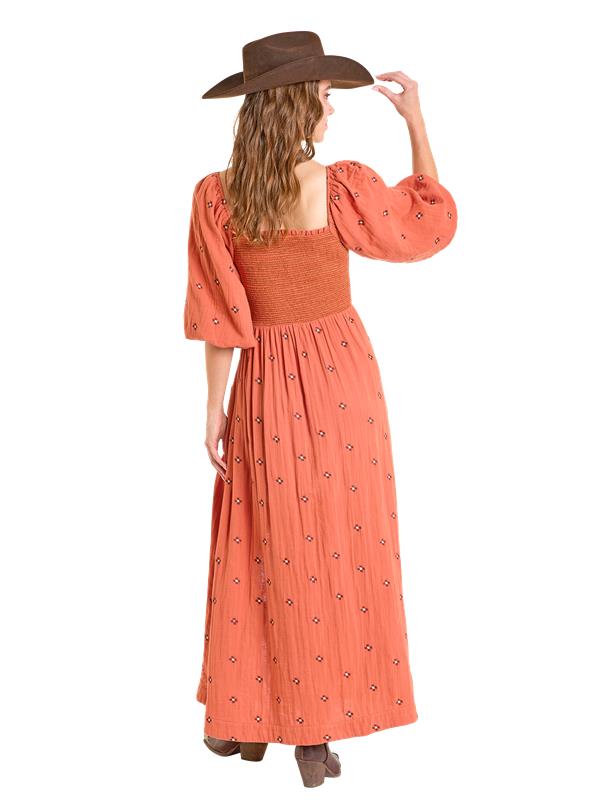 A woman wearing the Rock & Roll Denim Embroidered Maxi Dress stands facing away, holding a brown wide-brim hat. She has wavy hair and brown boots, showcasing the dress’s vibrant orange floral pattern and puffed sleeves.