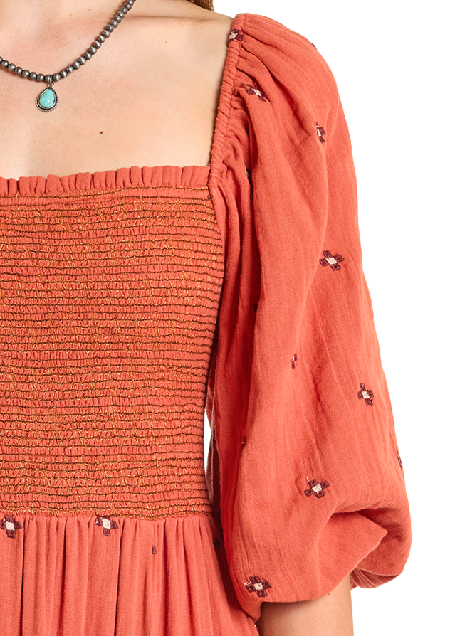 A woman wears the Embroidered Maxi Dress by Rock & Roll Denim, featuring coral smocked fabric, puffed sleeves, and small floral embroidery. She accessorizes with a silver necklace and turquoise pendant against a white background.