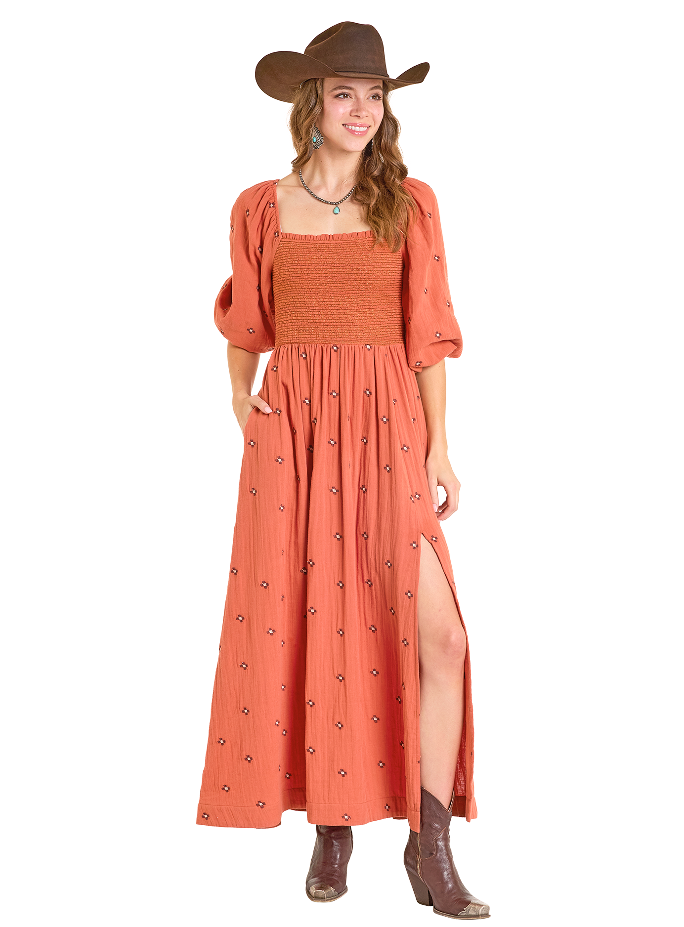 A woman in a Rock & Roll Denim Embroidered Maxi Dress, brown cowboy hat, ankle boots, and turquoise jewelry stands smiling with one hand in her pocket.