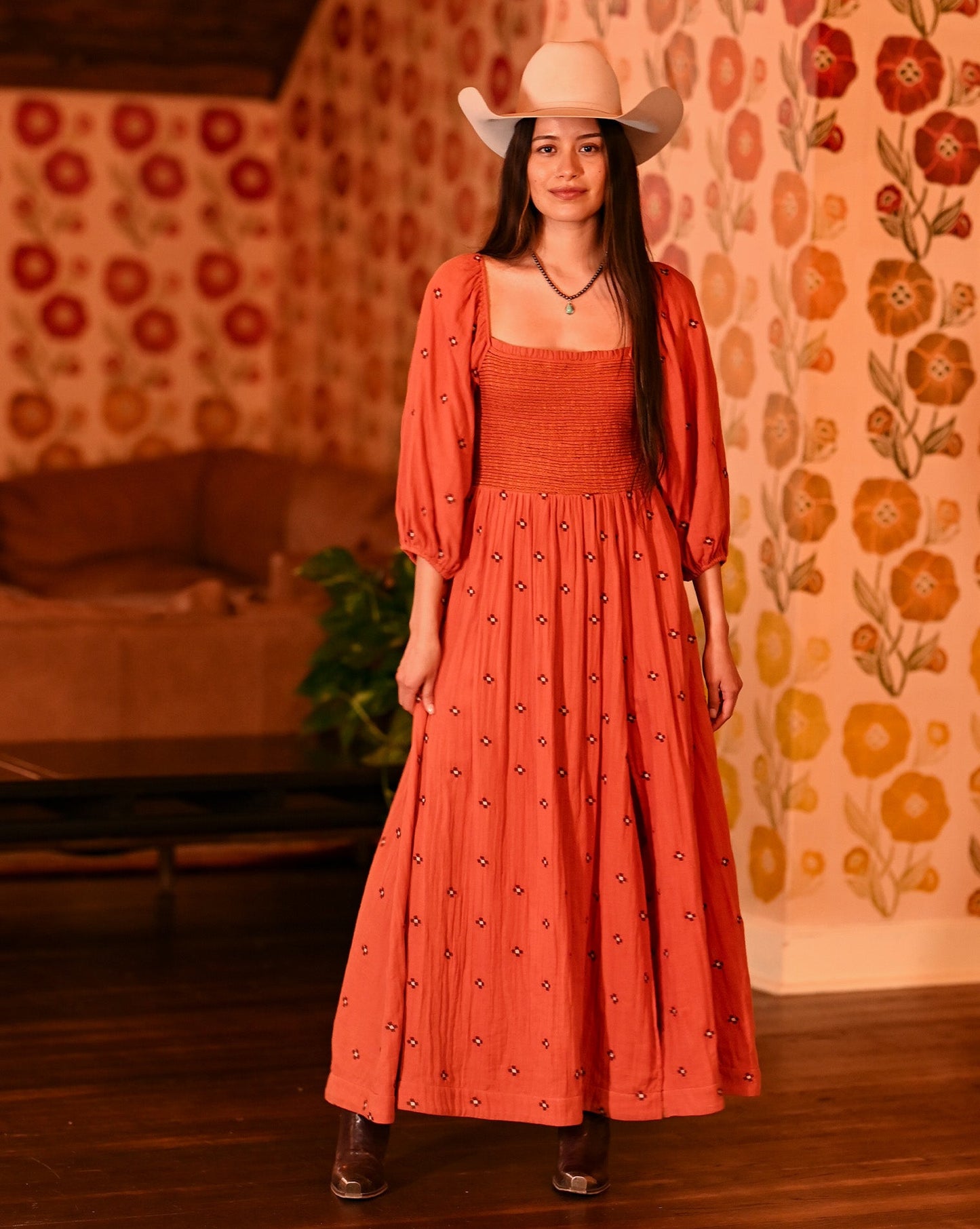A woman stands indoors on a wooden floor in front of floral wallpaper, wearing the Rock & Roll Denim Embroidered Maxi Dress and a white cowboy hat.