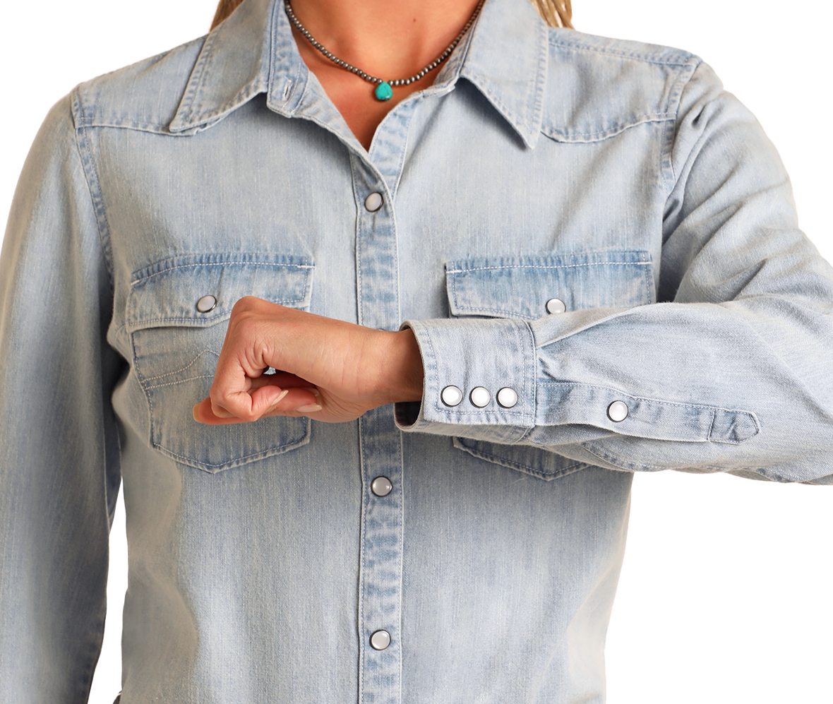 Wearing the Rock & Roll Denim Long Sleeve Snap Shirt and a turquoise necklace, a person poses with their right arm bent, closed fist on their left chest, evoking a subtle cowgirl vibe.