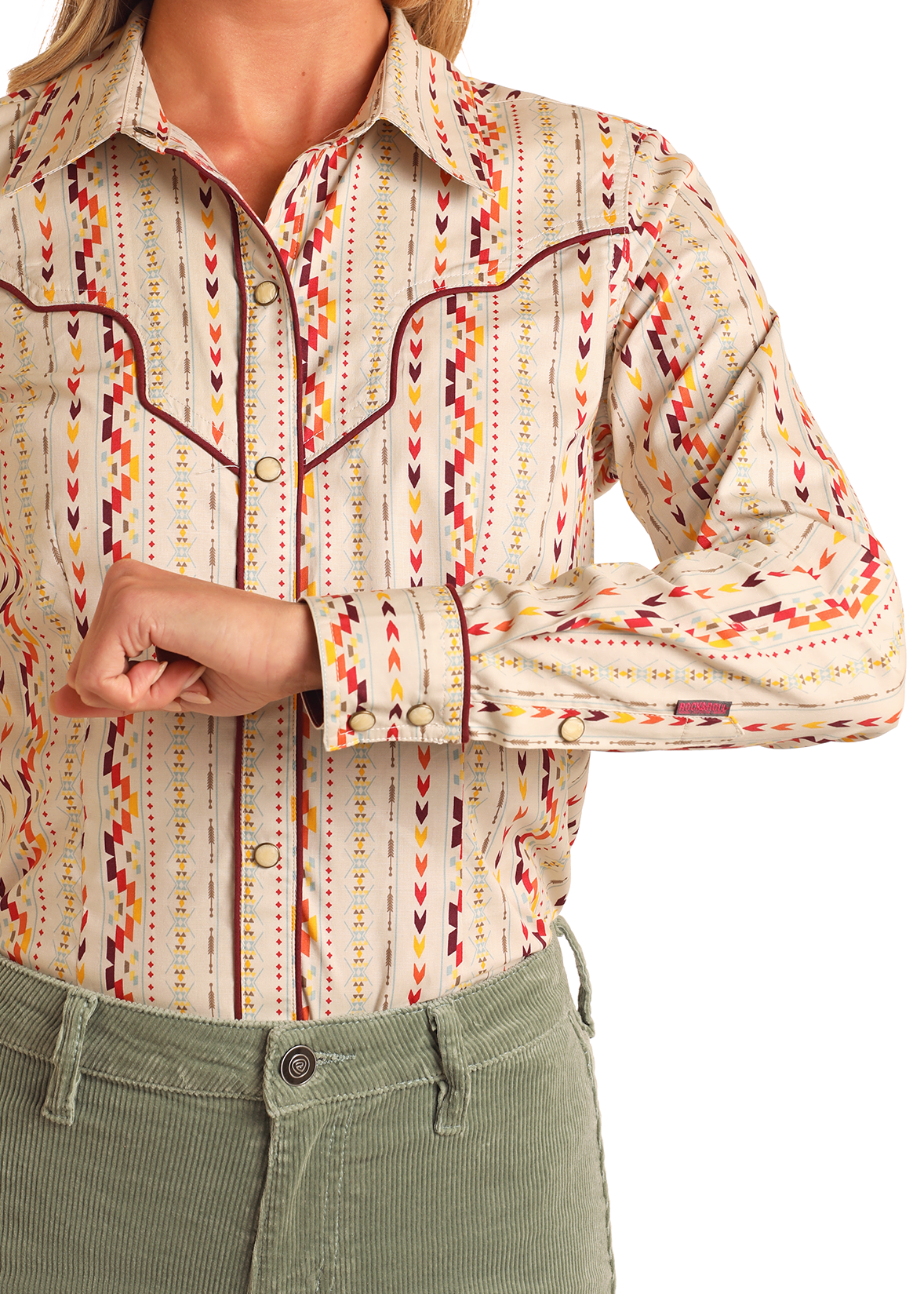 A person wears the Rock & Roll Denim Aztec Striped Long Sleeve Snap Shirt with red, yellow, and brown patterns, paired with light green corduroy pants. Their right arm is bent, hand near their left elbow.