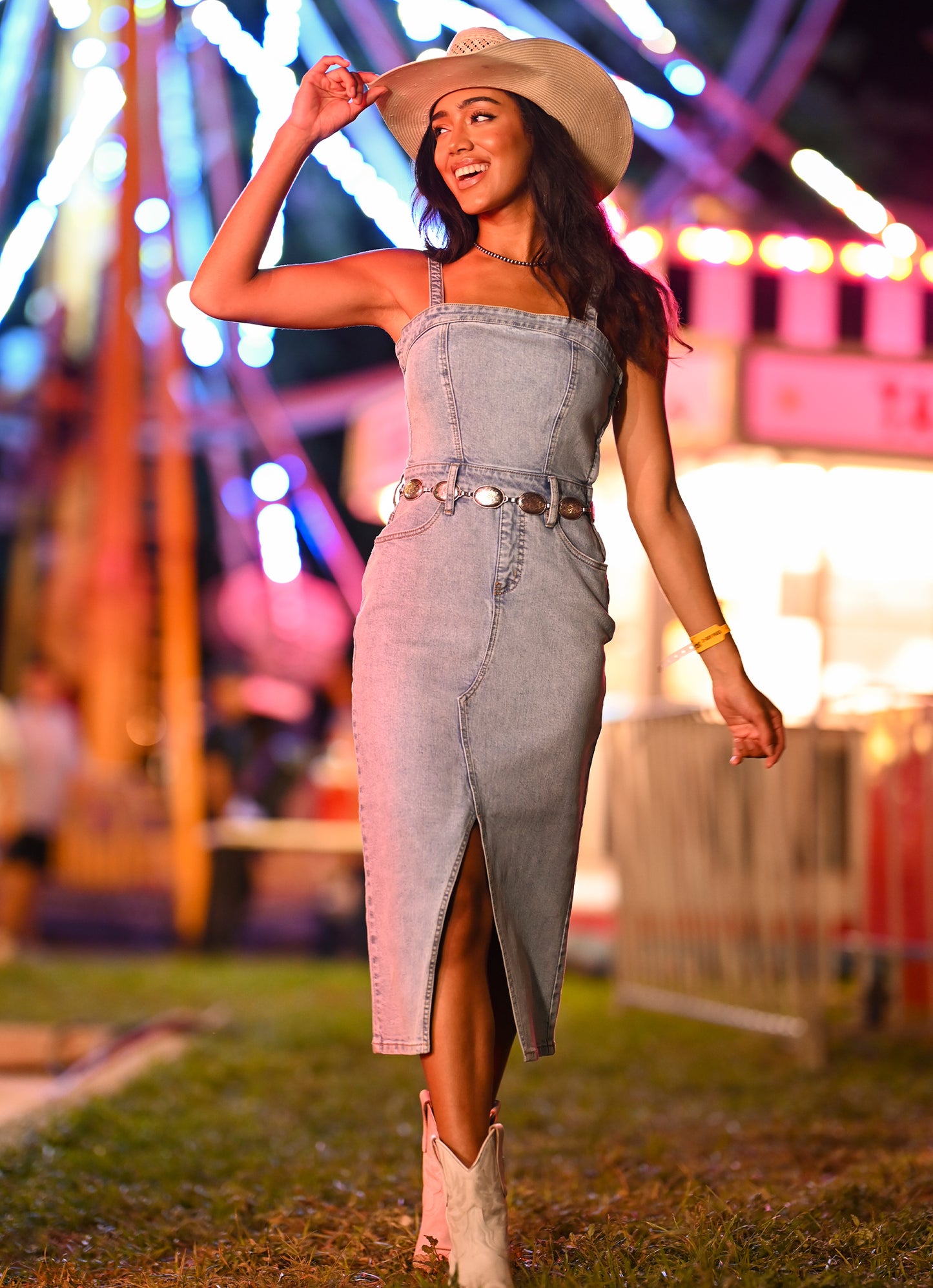 A woman wears the Rock & Roll Denim Detachable Strap Denim Midi Dress, paired with a cowboy hat and boots, smiling as she strolls outside at night with bright lights and a ferris wheel creating a festive backdrop.
