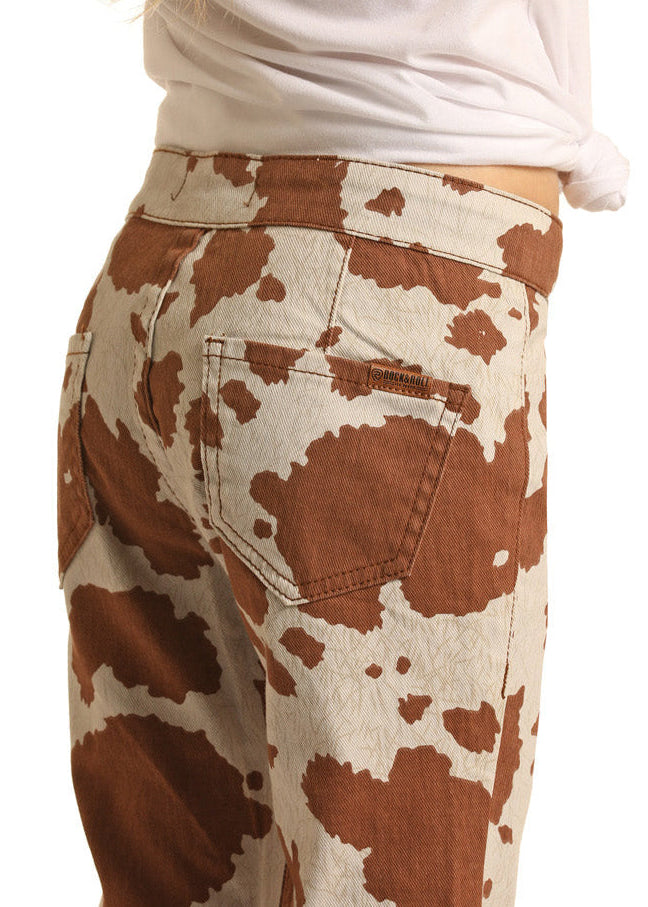 A little cowgirl wears Rock & Roll Denim Girls' Cow Print Bell Bottom Jeans in high-waisted brown and cream, paired with a white waist-tied shirt, shown from the back against a white background.