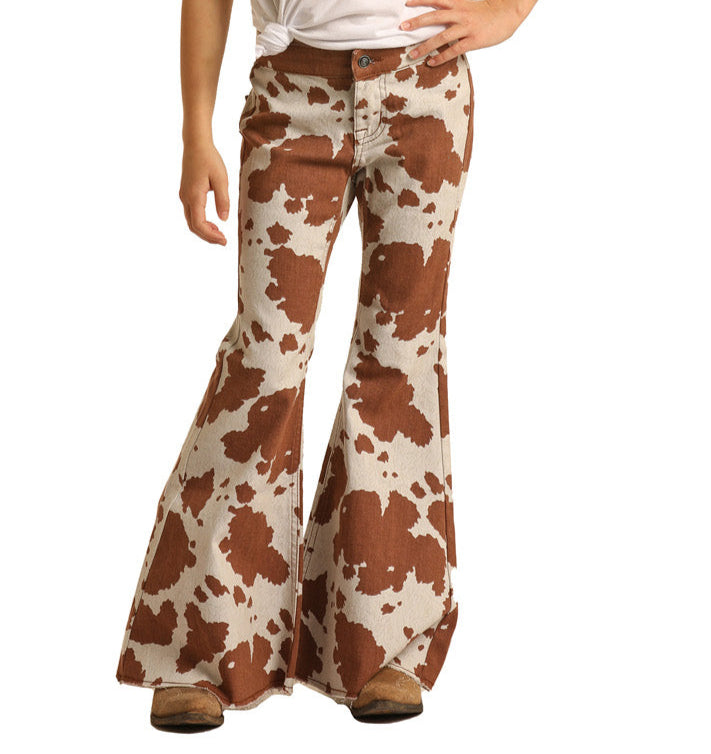 A little cowgirl stands confidently with one hand on her hip, wearing Rock & Roll Denim Girls' Cow Print Bell Bottom Jeans, a white shirt, and brown shoes.