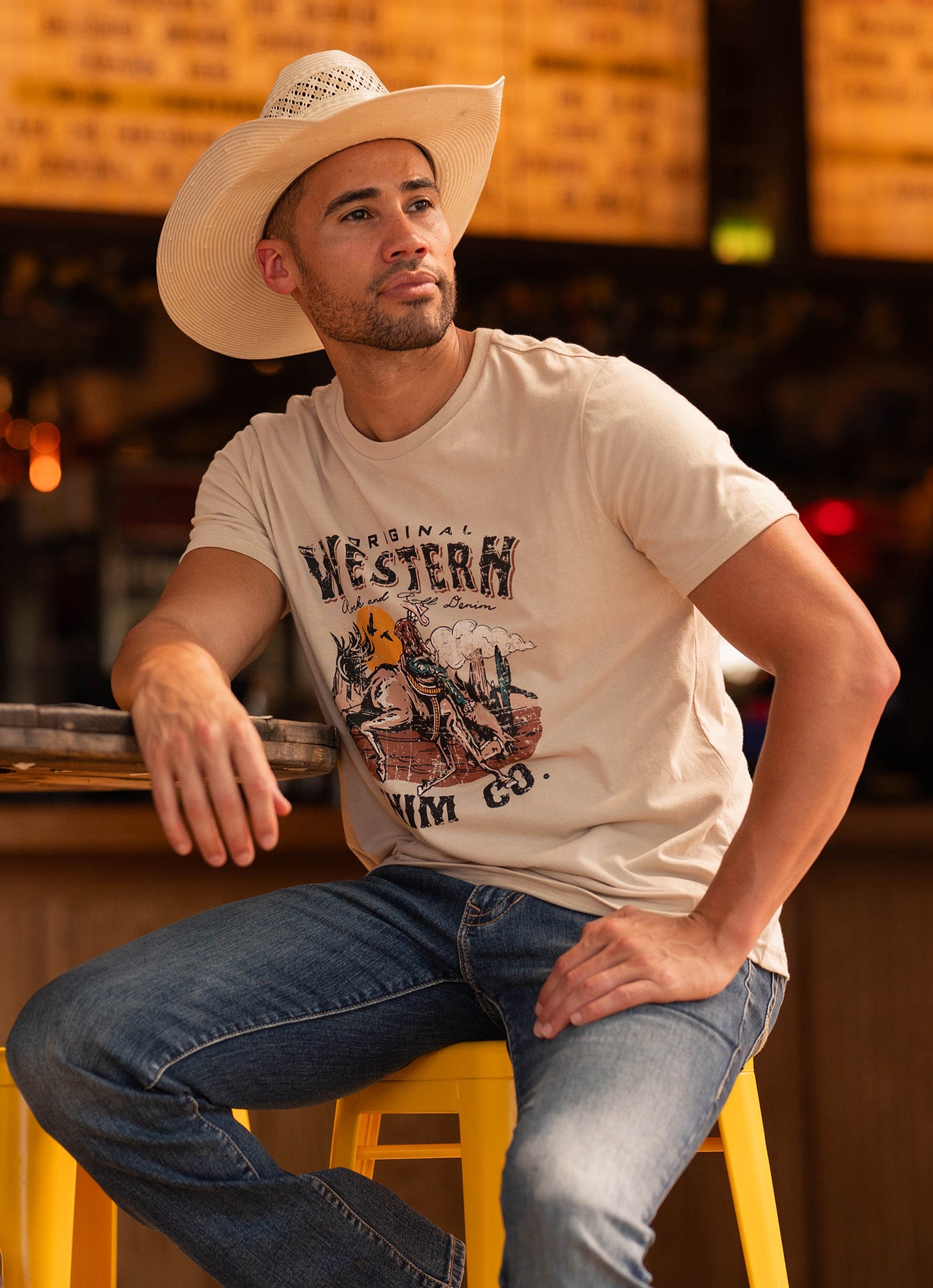 A man in a cowboy hat, Rock & Roll Denim's Original Western Denim Co. Graphic Tee, and jeans sits on a yellow stool indoors, gazing thoughtfully to the side.