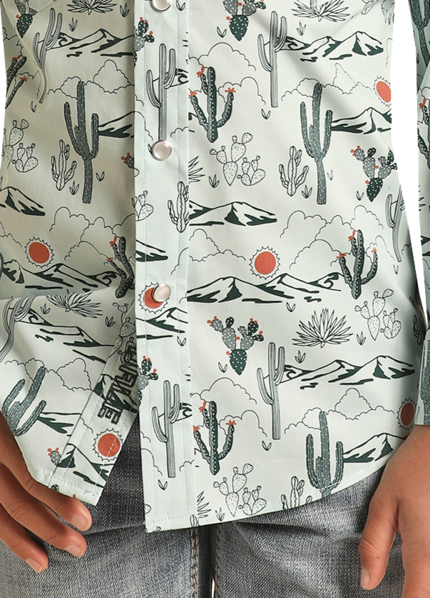 Close-up of a boy wearing the Rock & Roll Denim Boys' Dale Brisby Cactus Print Long Sleeve Snap Shirt featuring desert scenes; styled with light denim jeans, his hand rests by the buttons.