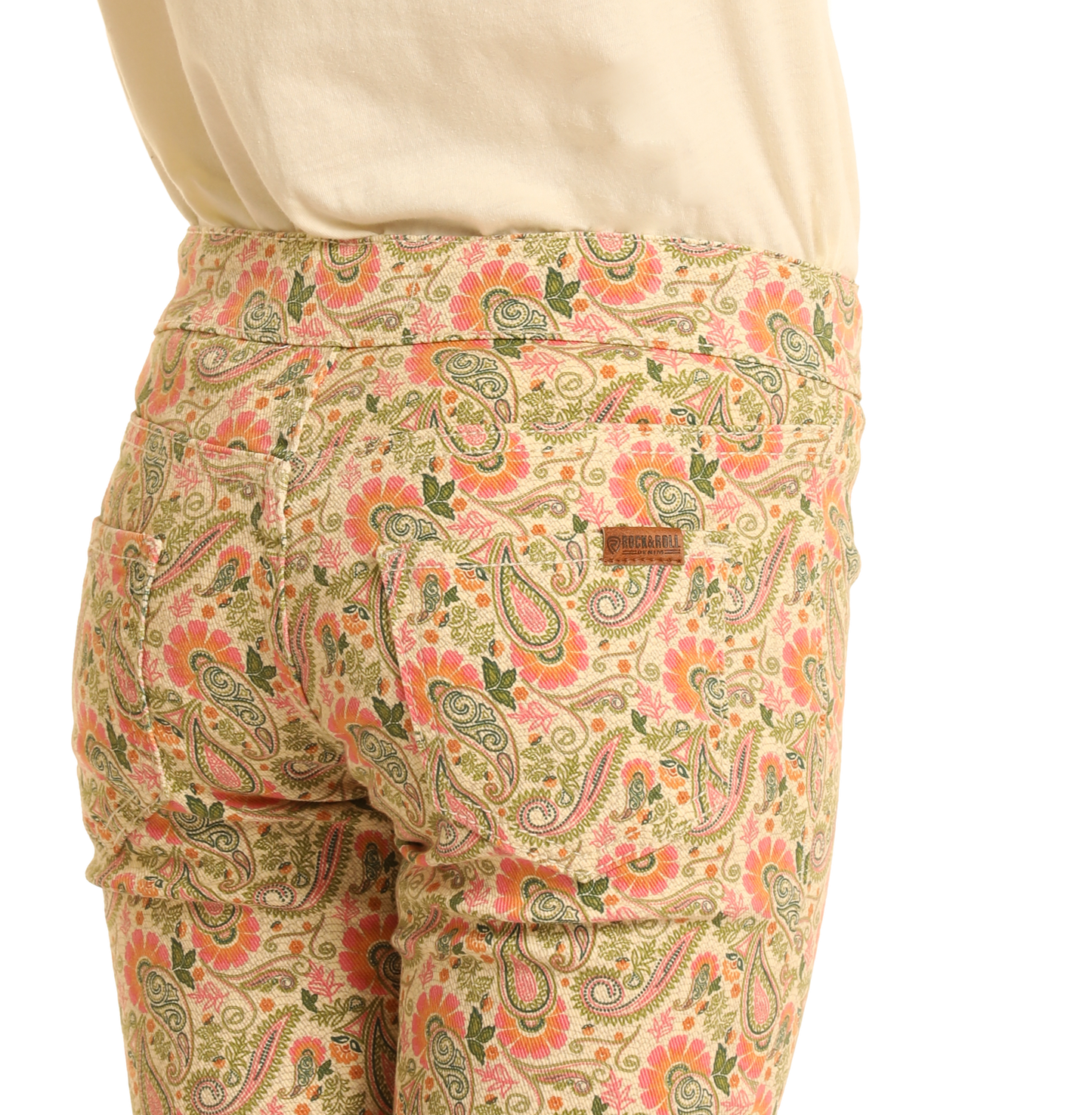 A person wears Rock & Roll Denim Girls' Paisley Print Flare Jeans in light beige with a colorful paisley pattern, paired with a cream top; the back pocket has a Dreamland Syndicate patch.