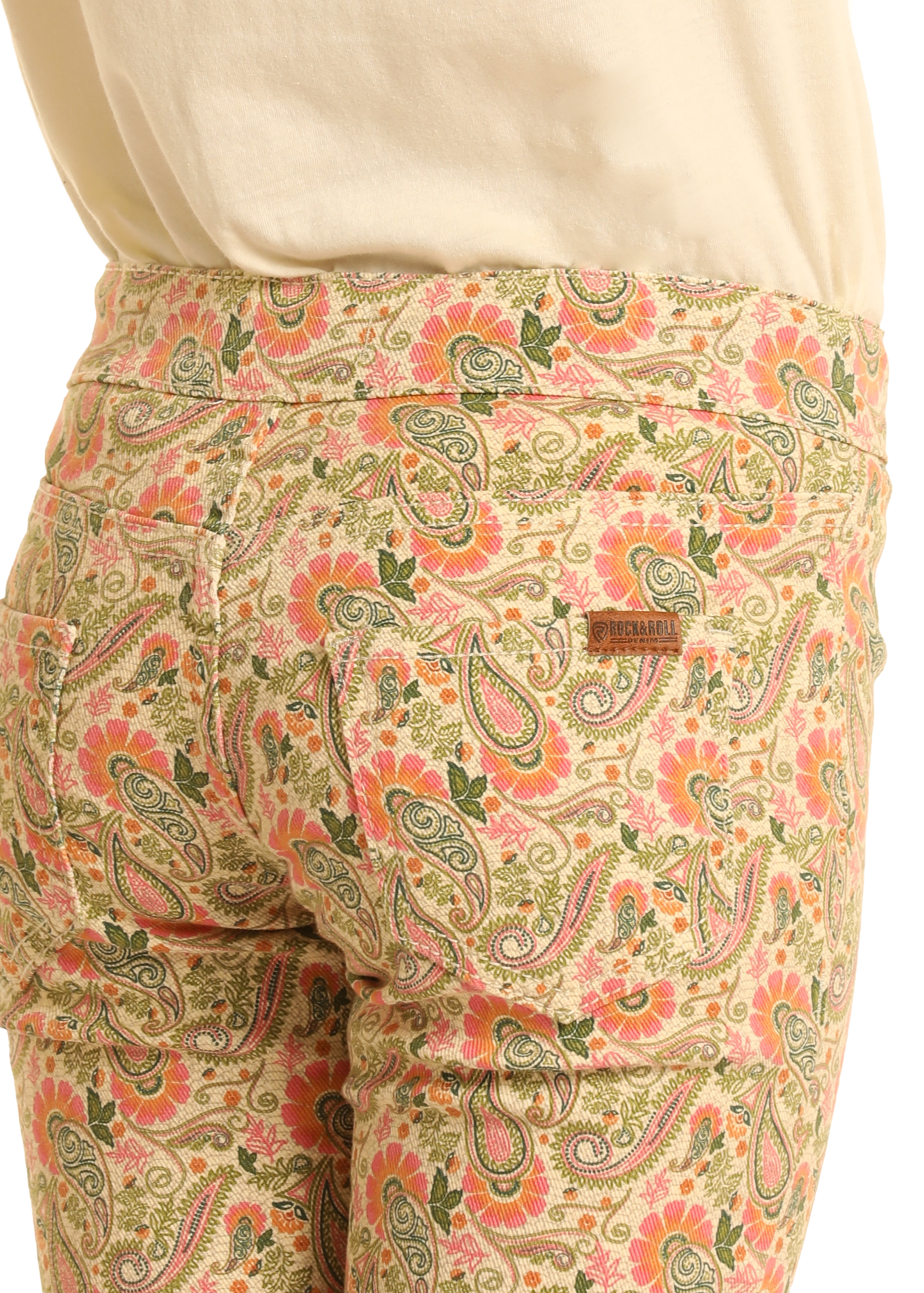 A person wears Rock & Roll Denim Girls' Paisley Print Flare Jeans in light beige with a colorful paisley pattern, paired with a cream top; the back pocket has a Dreamland Syndicate patch.