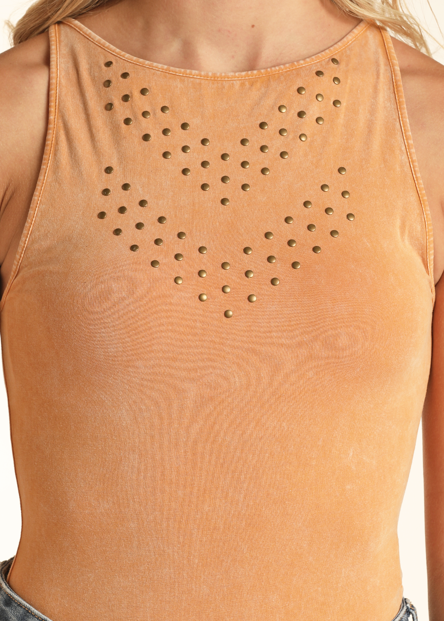 A close-up of a person wearing the Rock & Roll Denim Studded Bodysuit, a sleeveless mineral wash orange piece with gold studs in curved lines on the chest, tucked into light blue jeans.