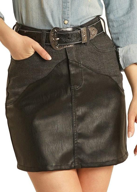 A person wears a light denim shirt with rolled-up sleeves, paired with the Rock & Roll Denim Black Western Yoke Mini Skirt featuring pockets and a black belt with a silver buckle. Their left hand rests in a pocket, embodying the brand's signature style.