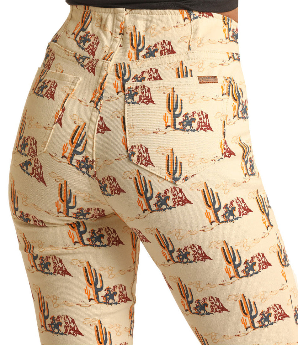 A person is shown from the back waist down wearing Rock & Roll Denim’s Desert High Rise Pull-On Flare Jeans in beige with a colorful southwestern desert print, paired with a black top.