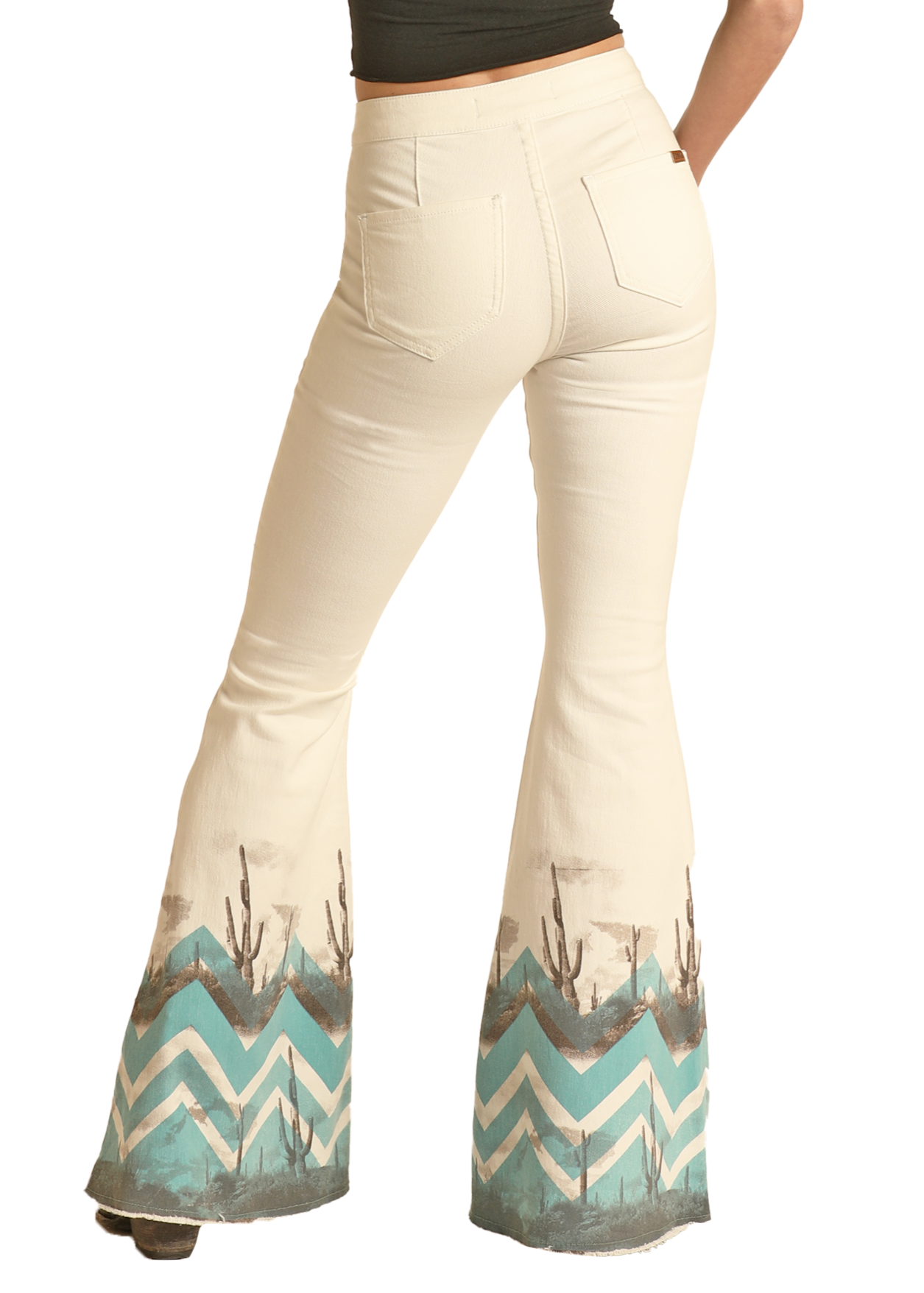 A person models the Chevron Desert Print High Rise Bell Bottom Jeans by Rock & Roll Denim, featuring cream fabric, teal chevron desert print, and brown cactus accents near the hem, paired with a dark crop top for western flair.