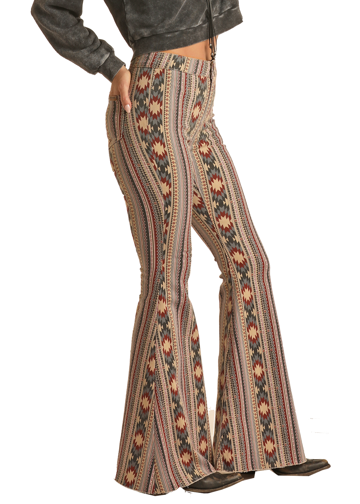 A person stands in profile, hands on hips, wearing Rock & Roll Denim’s Allover Aztec High Rise Bell Bottom Jeans with a geometric pattern, paired with a cropped long-sleeve sweater and black boots.