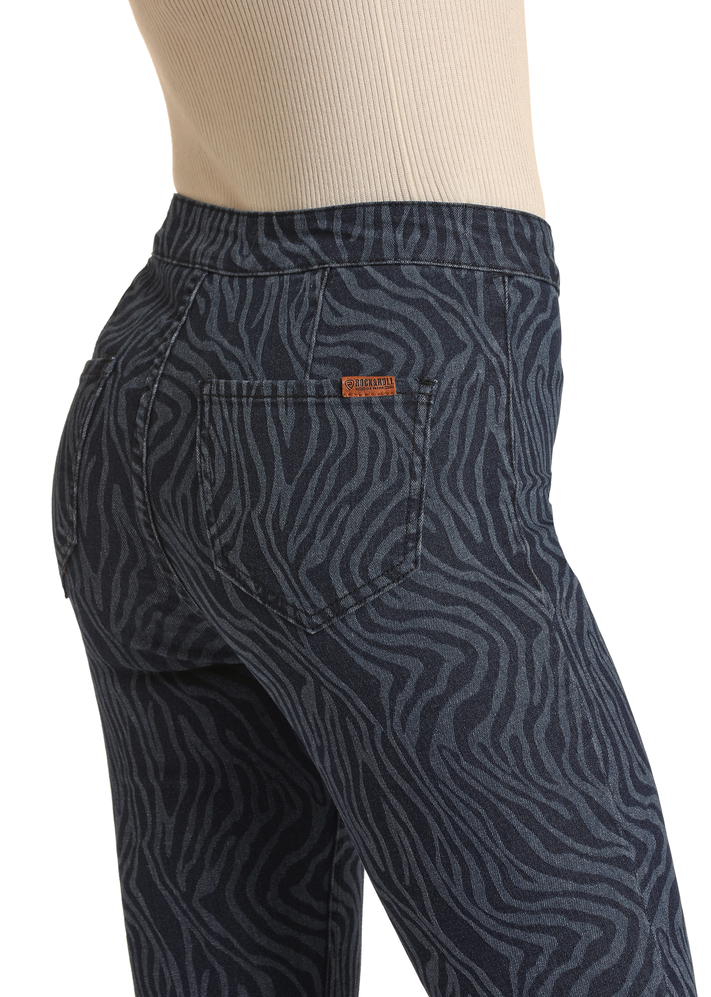 The image, cropped from the waist down, shows a person wearing Rock & Roll Denim Zebra High Rise Bell Bottom Jeans in dark blue with a black zebra print pattern and a small brown leather patch on the back pocket, paired with a beige ribbed top.