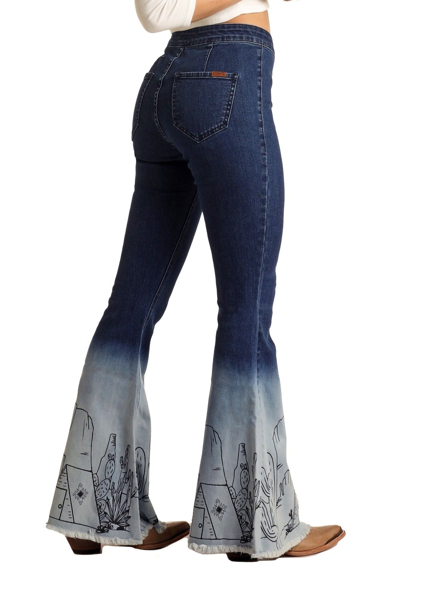 A person wearing Rock & Roll Denim Desert Ombre High Rise Bell Bottom Jeans featuring dark blue to faded light blue ombre and cactus and cowboy boot embroidery near the hem, styled with brown heeled boots for southwestern flair.