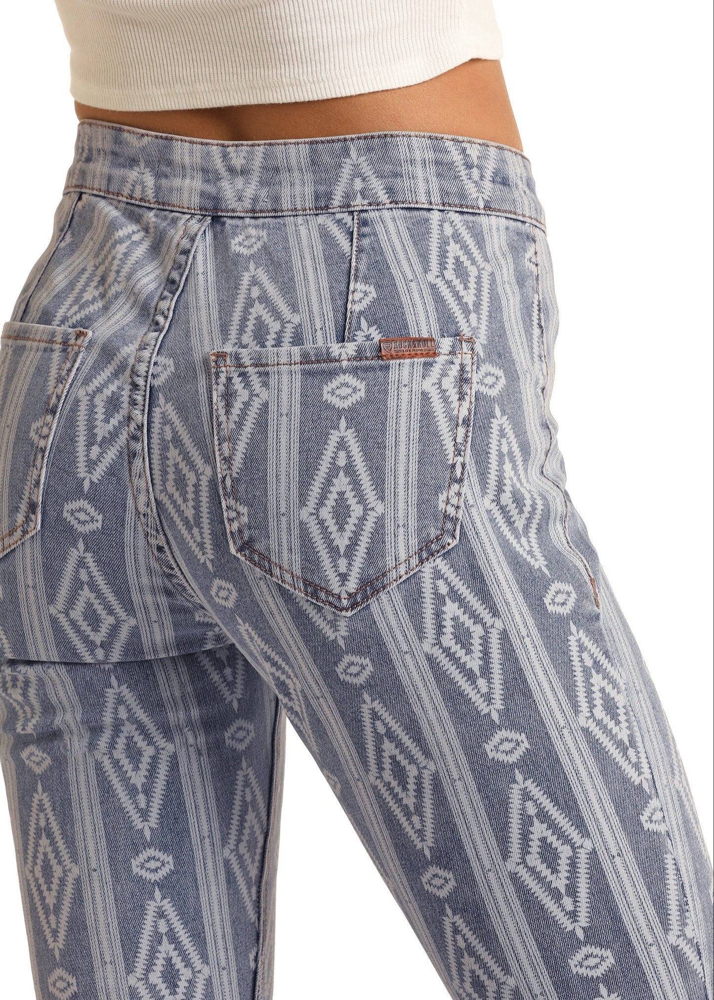 A person wears Rock & Roll Denim Aztec High Rise Bell Bottoms in light blue with a white geometric pattern, paired with a white ribbed crop top. The image highlights the unique Aztec patterned jeans and standout back pockets.