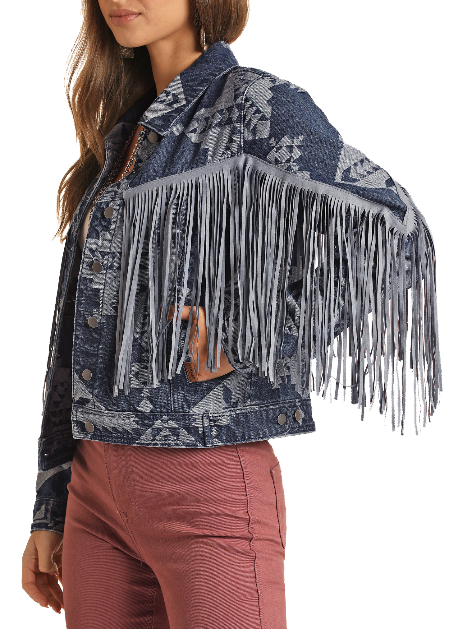 A woman models the Rock & Roll Denim Fringe Aztec Print Denim Jacket, featuring bold patterns and long gray fringe on the sleeves, paired with mauve pants. She poses with one arm raised to highlight the unique fringe detail.