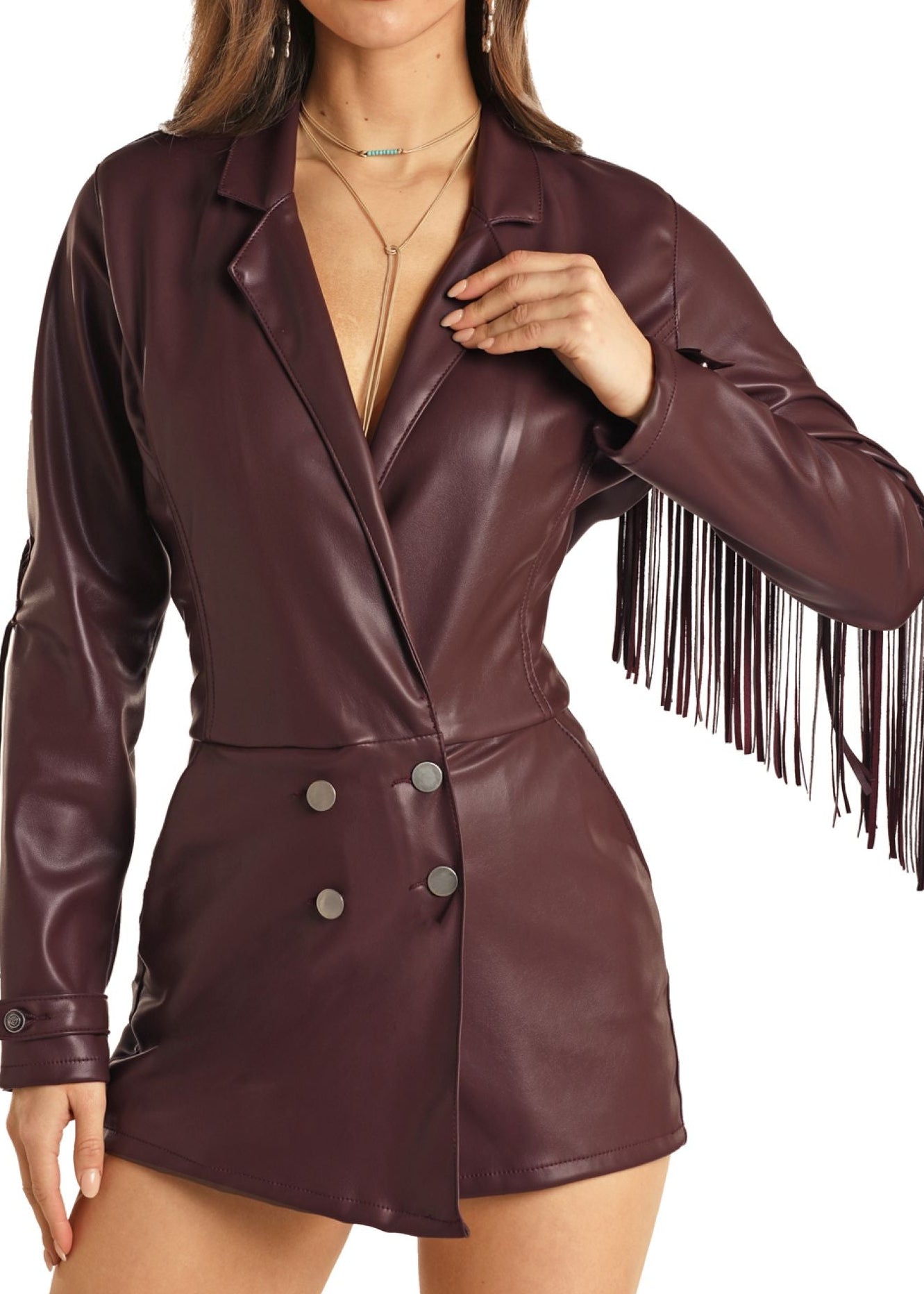 A woman models the Rock & Roll Denim Fringe Pleather Romper in burgundy featuring long sleeves, fringe on one sleeve, and double-breasted buttons. She wears layered necklaces with her long hair; her head is not shown.
