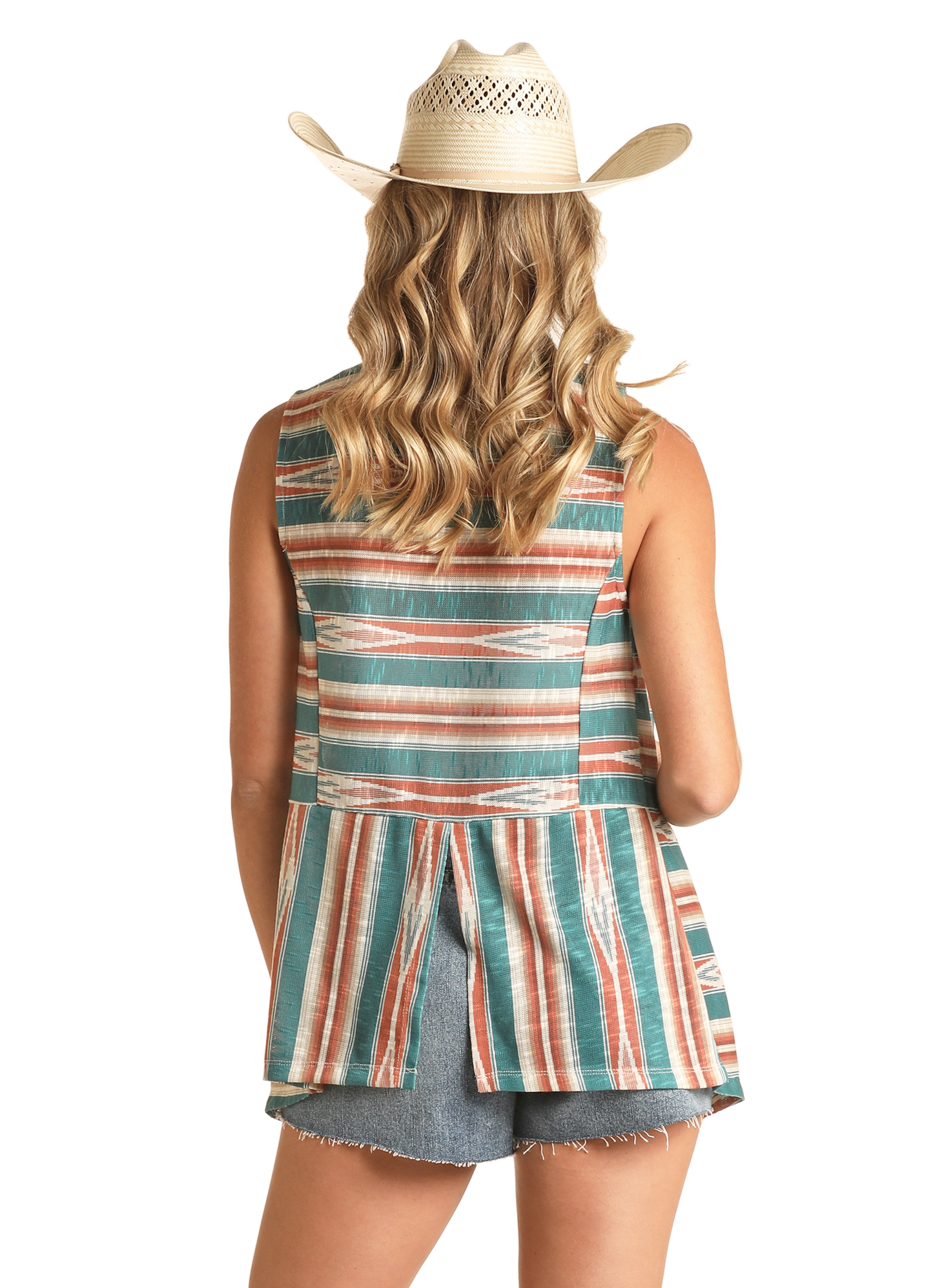 A woman with wavy blonde hair wears a Rock & Roll Denim Turquoise Aztec Drape Front Vest, paired with a straw cowboy hat, sleeveless striped top, and frayed denim shorts, standing with her back to the camera against a plain background.