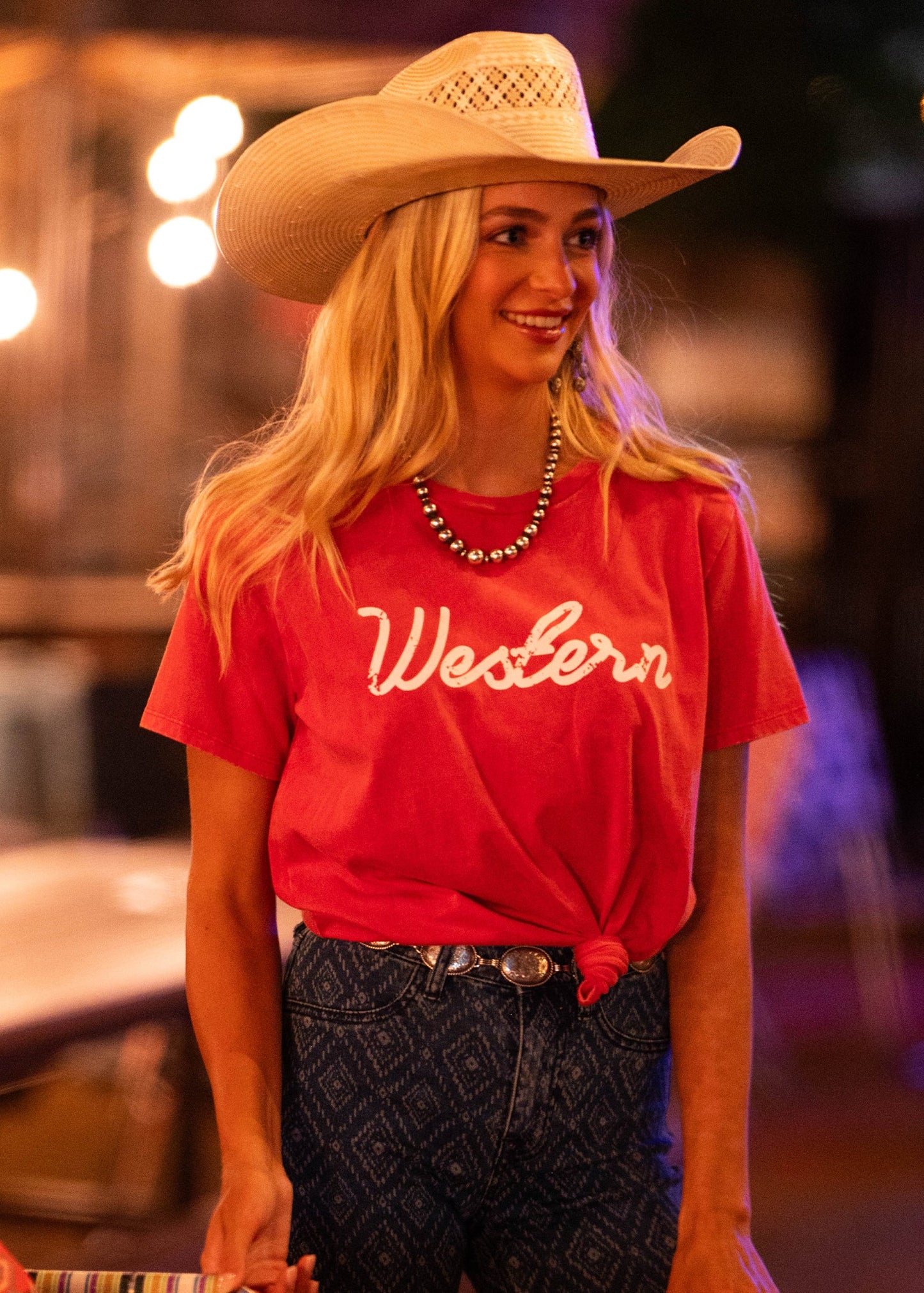 A smiling woman in a cowboy hat and long blonde hair stands indoors, wearing the Rock & Roll Denim Western Graphic Tee with a necklace and patterned jeans. The warmly lit, softly blurred background highlights her casual style.