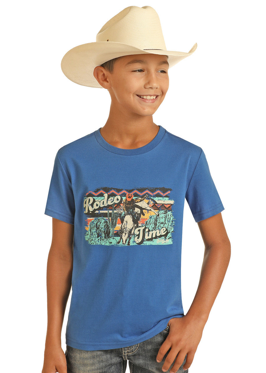 A smiling boy in a cowboy hat wears the Rock & Roll Denim Boys' Dale Brisby Rodeo Time Graphic T-Shirt, standing with one hand in his pocket against a plain white background.