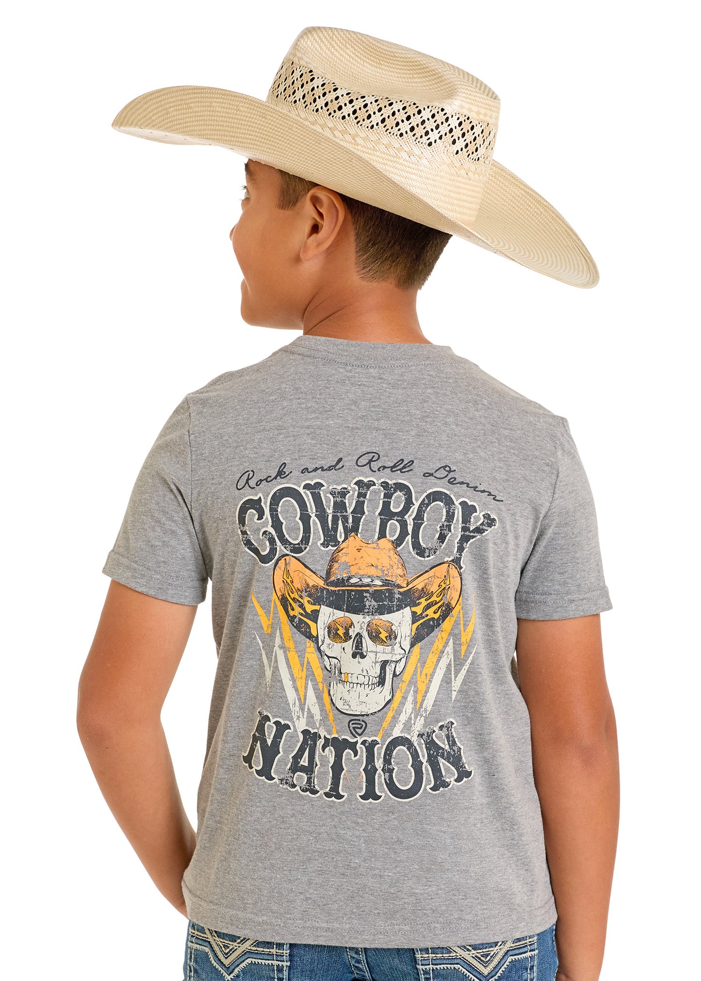 A boy in a straw cowboy hat and the Rock & Roll Denim Boys' Cowboy Skull Graphic Tee, featuring a bold skull, cowboy hat, and lightning design on the back, stands facing away, embodying adventurous style.