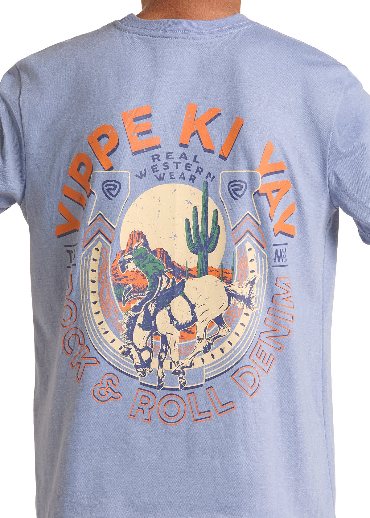A close-up of a light blue t-shirt showing the back graphic. The graphic features a large western-themed design with a cowboy on a bucking horse inside a horseshoe, a green saguaro cactus, and orange text reading 'VIPPE KI YAK' and 'REAL WESTERN WEAR'.
