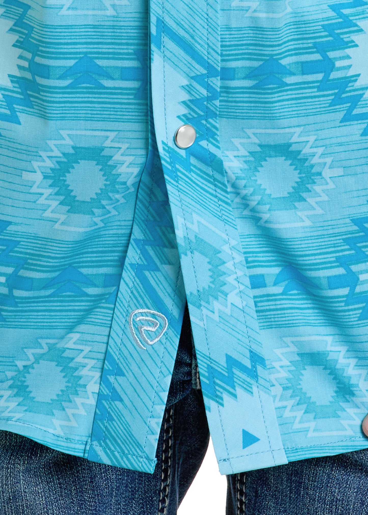 Close-up of the Rock & Roll Denim Boys' Turquoise Aztec Short Sleeve Snap Shirt (M) featuring a geometric pattern, silver snap buttons, and a small logo near the hem, paired with blue denim jeans.