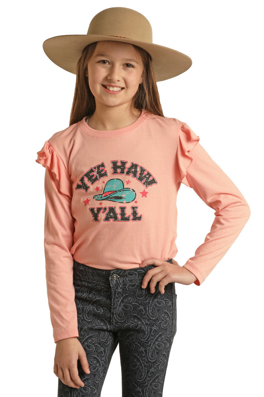 A smiling girl in a wide-brimmed tan hat and dark paisley pants wears the Rock & Roll Denim Girls' Yee Haw Y'all Graphic T-Shirt with cool ruffles and a cowboy hat graphic, posing confidently and radiating Southern charm.