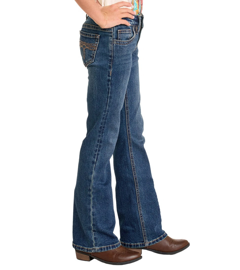 A person stands sideways wearing Rock & Roll Denim Girls' Boot Stitch Bootcut Jeans, brown cowboy boots, and a colorful shirt partly visible with hands on hips.