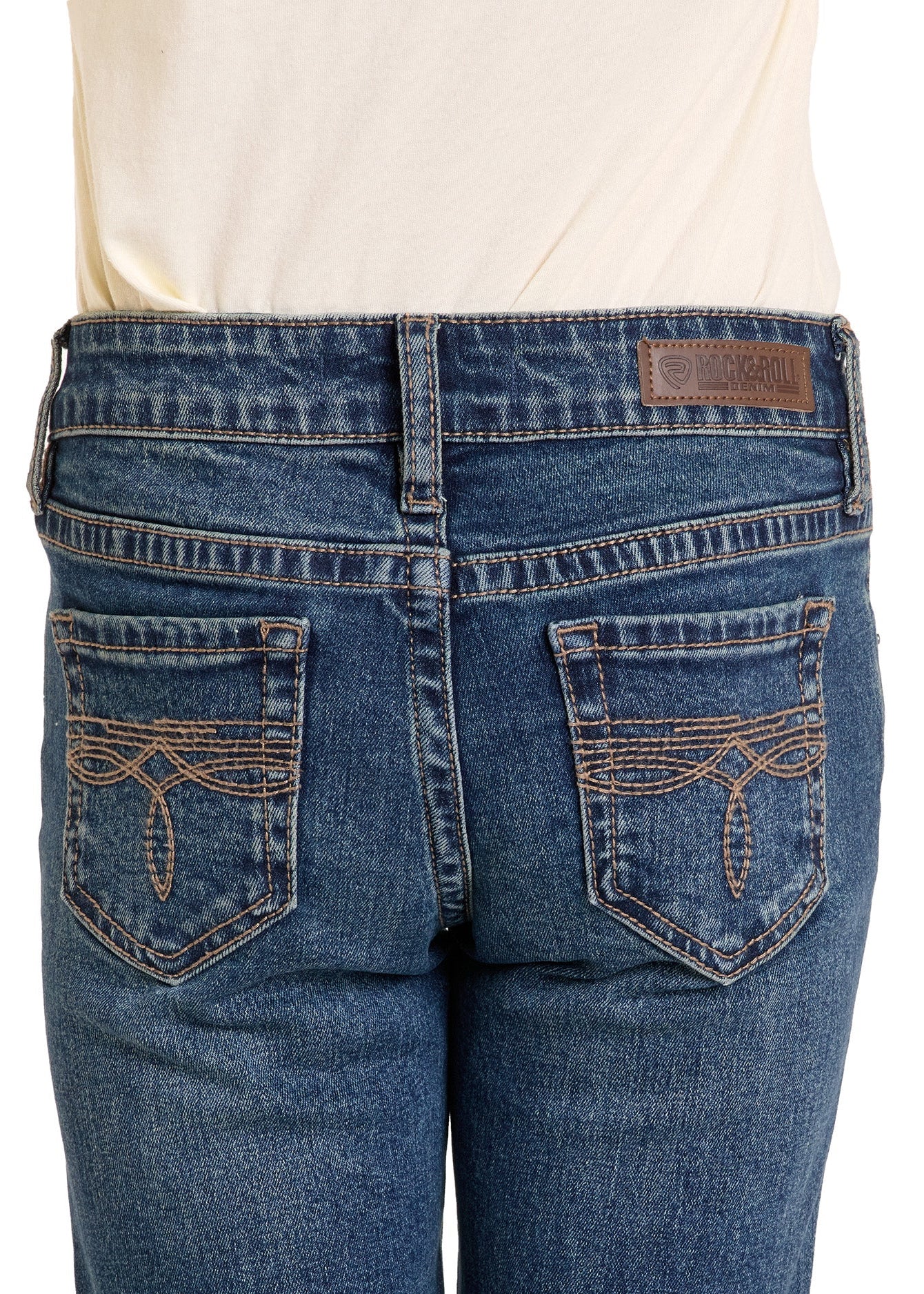 Close-up of Rock & Roll Denim Girls' Boot Stitch Bootcut Jeans, featuring decorative pocket stitching and a brown leather patch on the waistband, worn with a cream-colored shirt.