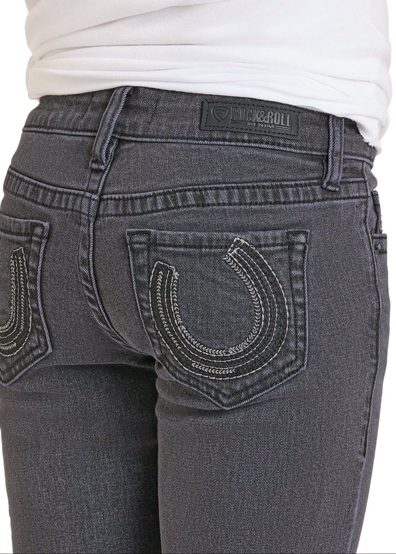 A girl wears Rock & Roll Denim’s Extra Stretch Horseshoe Bootcut Jeans in dark gray with white-stitched back pockets and a black brand patch, paired with a white top for a Western-inspired look.