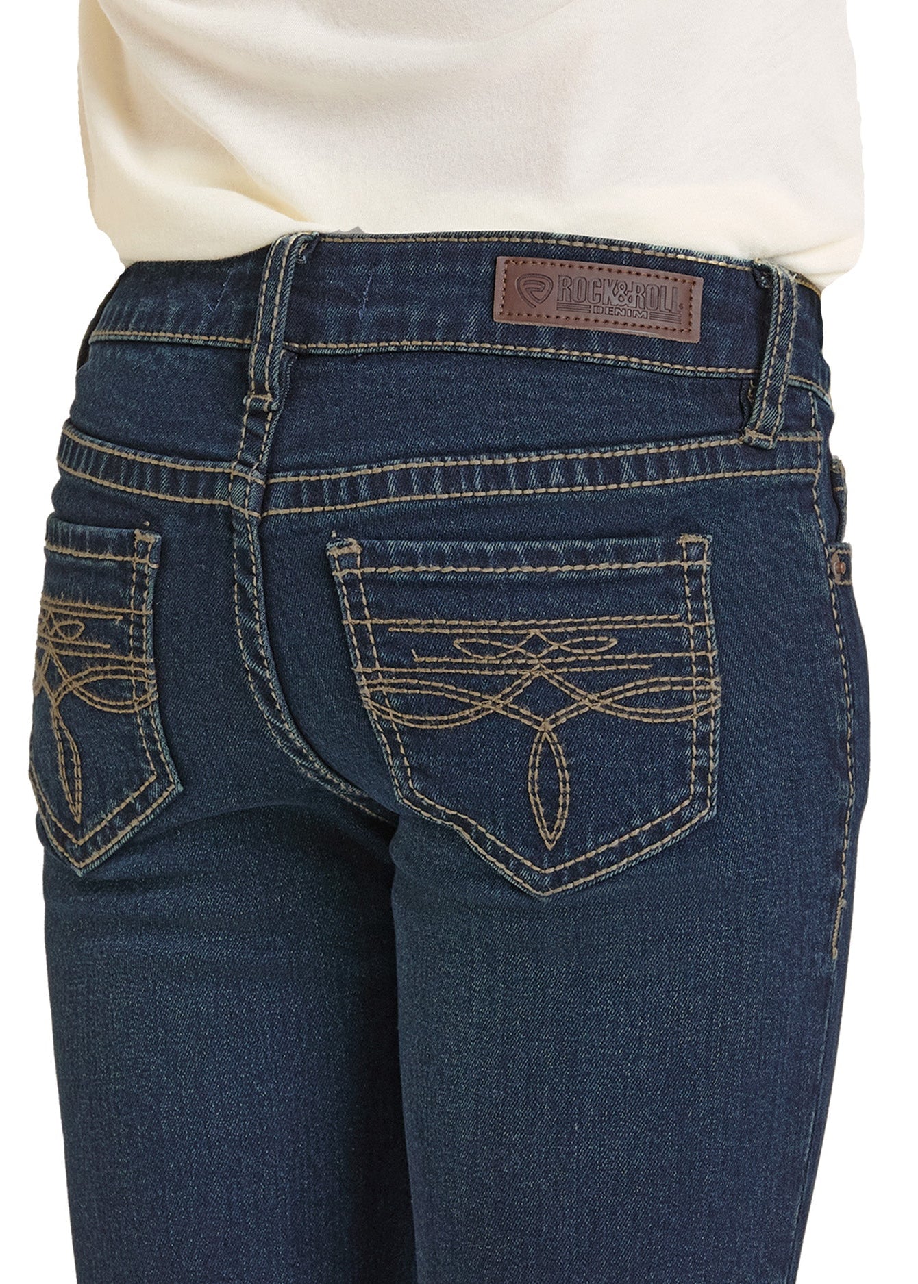 Close-up of a person in Rock & Roll Denim Girls' Dark Wash Boot Stitch Bootcut Jeans featuring beige stitching on the back pockets, a brown leather brand patch on the waistband, and a white top.