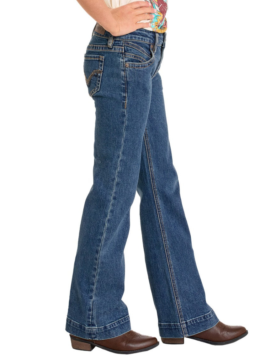 A girl stands in profile against a white background, wearing Rock & Roll Denim Girls' Signature Stitch Trouser Jeans and brown cowboy boots, with one hand resting on her hip.