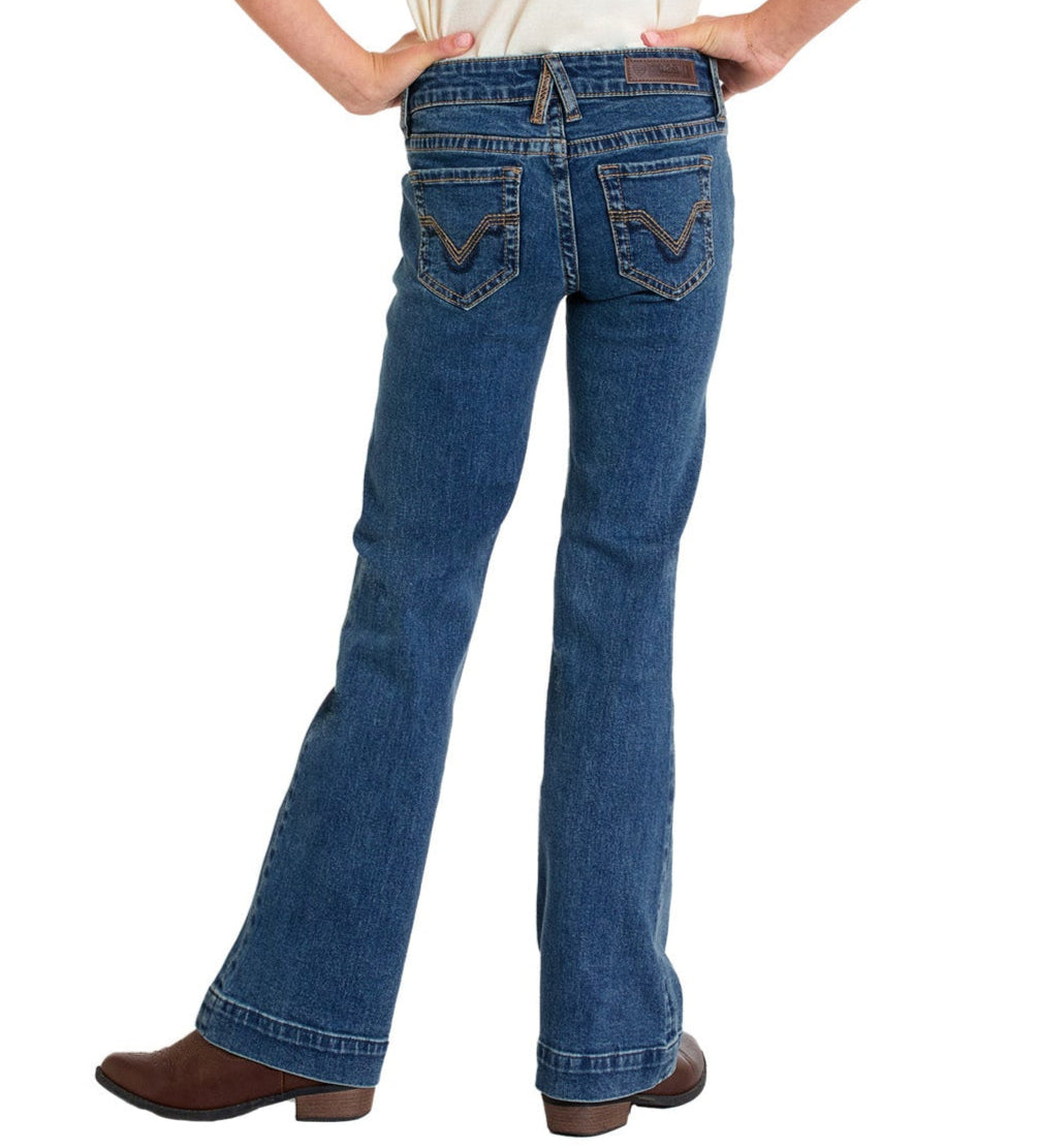 A girl wearing Rock & Roll Denim Girls' Signature Stitch Trouser Jeans stands with hands on hips, facing away from the camera; the blue jeans feature decorative back pocket stitching.