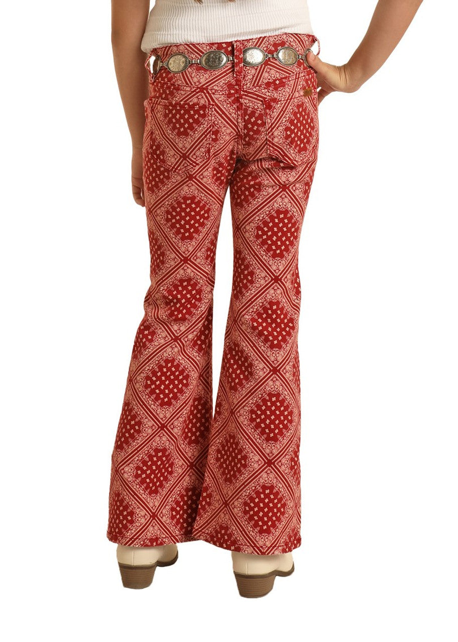 A person wears Rock & Roll Denim Girls' Bandana Print Flare Jeans with western flair, a white top, decorative silver belt, and tan heeled shoes, shown from the back with one hand on their hip.