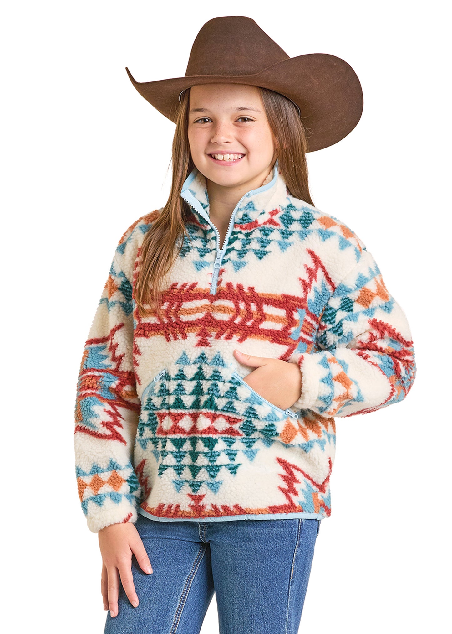 A smiling girl wears the Girls’ Aztec Sherpa Pullover by Rock & Roll Denim, featuring plush red, teal, and orange designs, a brown cowboy hat, and blue jeans. She stands with one hand in her pocket against a white background.
