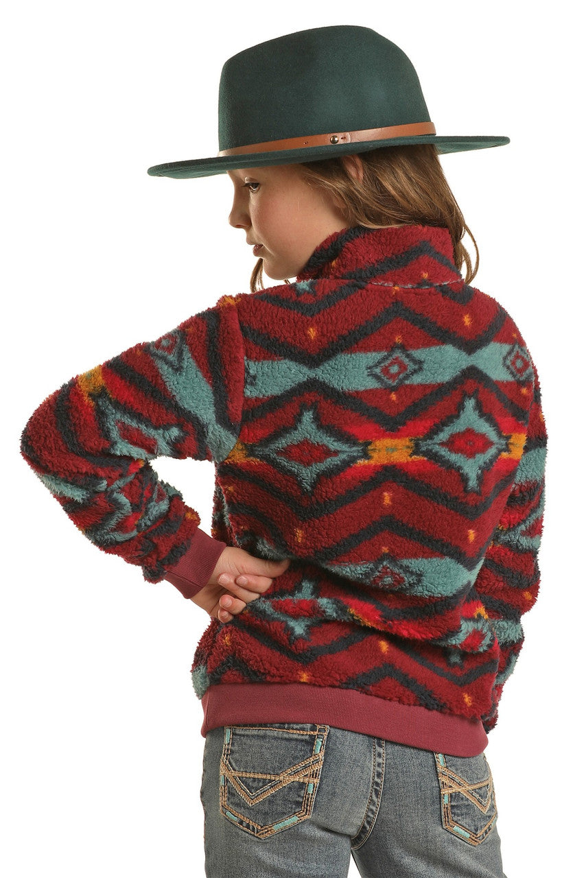 A child stands with hand on hip, facing away, wearing a green wide-brimmed hat, blue jeans with decorative stitching, and a Rock & Roll Denim Girls' Aztec Print Pullover in vibrant red, teal, and yellow.