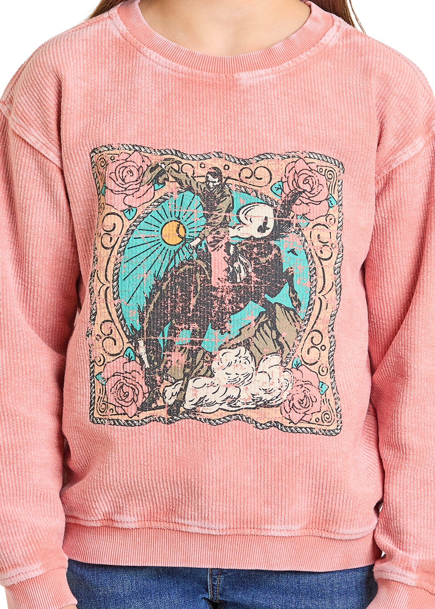A girl wears the Rock & Roll Denim Girls' Saddle Bronc Graphic Pullover, featuring a pink textured design with a vintage cowboy on horseback, surrounded by decorative patterns, roses, and a sun.