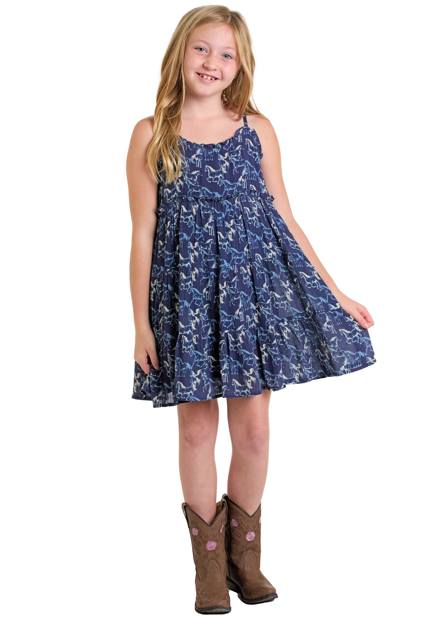 Smiling young girl with long blonde hair wears the Rock & Roll Denim Girls' Horse Dress and brown cowboy boots, standing on a white background and holding out one side of her sleeveless blue cowgirl dress.