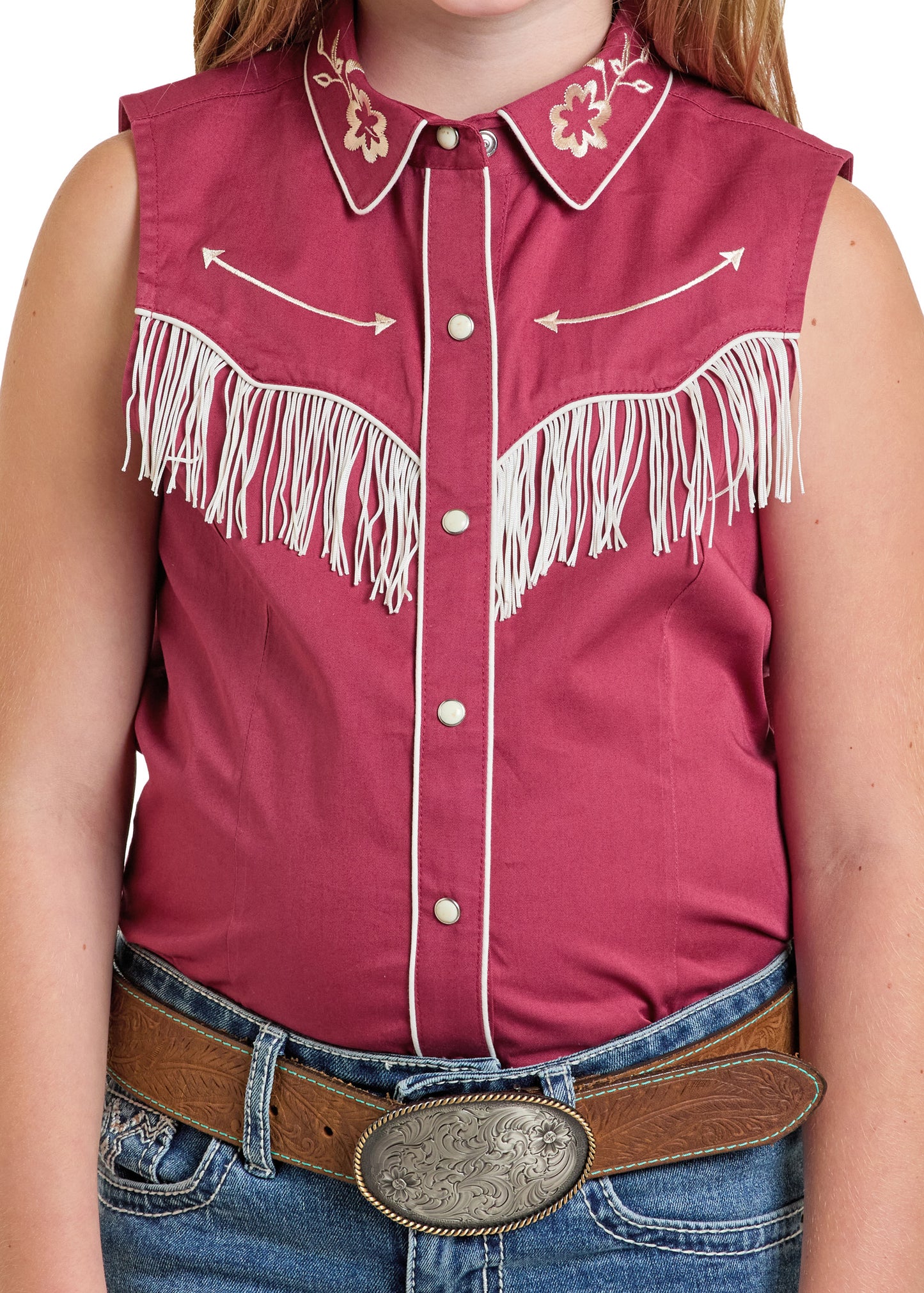 A girl wears a Rock & Roll Denim Girls' Retro Snap Shirt—sleeveless, maroon with white fringe, floral embroidery, and arrow designs—paired with blue jeans and a brown belt with an ornate silver buckle.