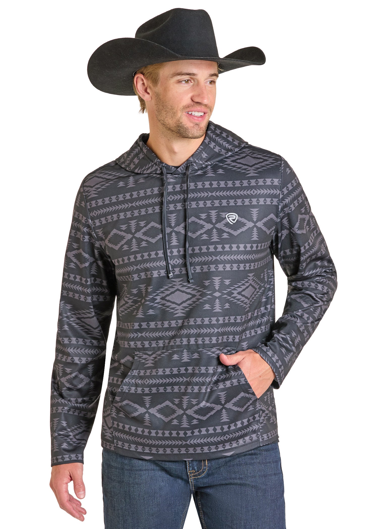 A man in a black cowboy hat, Rock & Roll Denim Black Aztec Performance Hoodie, and blue jeans stands angled to the side, smiling with one hand in his hoodie pocket—showcasing comfort and style.
