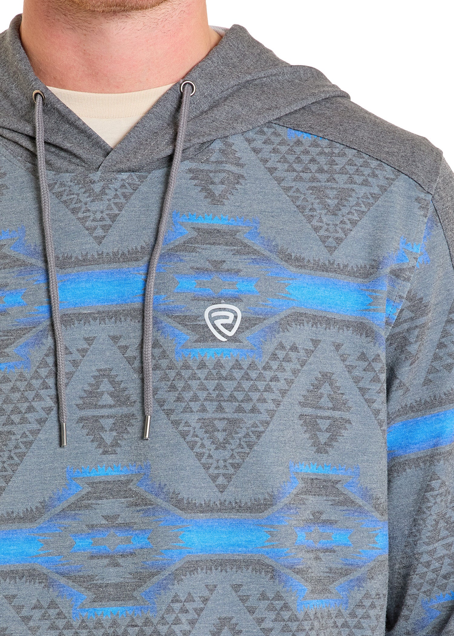 A person wears a Grey Aztec Performance Hoodie by Rock & Roll Denim, featuring blue and black geometric patterns, a small white logo on the chest, drawstrings, and their face partially out of frame.
