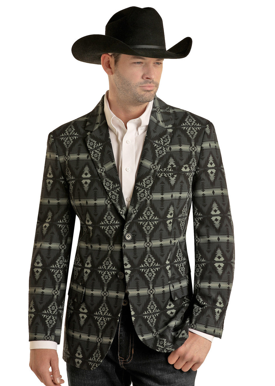 A man in a black cowboy hat, white dress shirt, and a Rock & Roll Denim Aztec Print Sport Coat with bold geometric designs stands with one hand by his side and the other in his pocket.